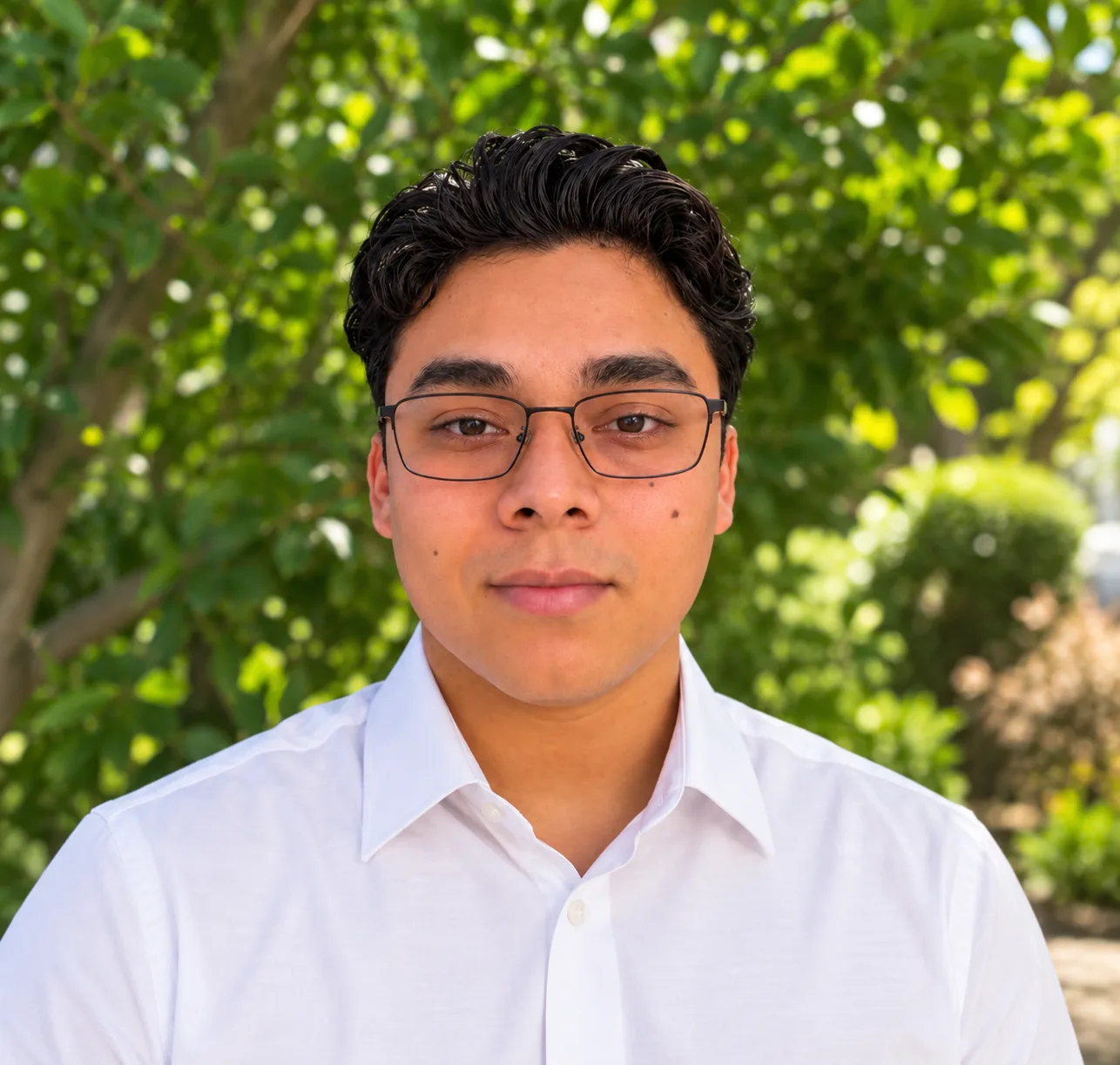 A young man with short dark hair and glasses wearing a white button-up shirt, standing outdoors in front of green leafy trees.